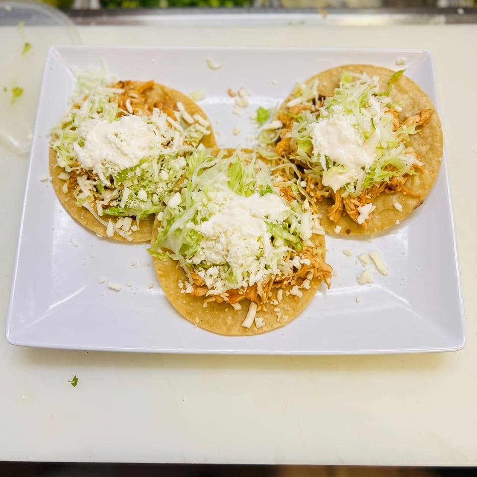 Three chicken tostadas topped with shredded lettuce, crumbled cheese, and sour cream on a white plate.