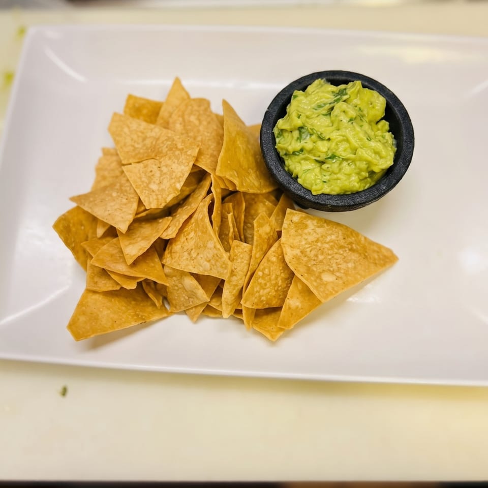 A white rectangular plate holds a pile of golden tortilla chips and a black bowl of guacamole.